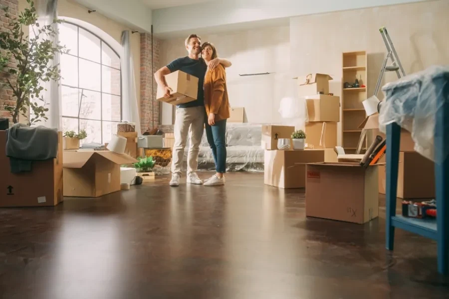 Casal jovem abraçado no meio de uma sala nova, rodeado por caixas de mudança, simbolizando a compra da sua primeira casa com apoio financeiro.